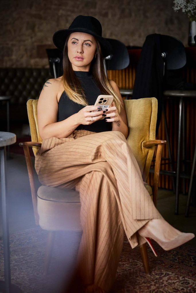 Personal Branding Business-Porträt: Eine Person in einer dunkleren Umgebung mit warmen Braun- und Schwarztönen. Freundlicher Ausdruck unterstreicht Professionalität und Persönlichkeit. Nachdenklicher Blick nach draußen, Person sitz und hat Handy in der Hand